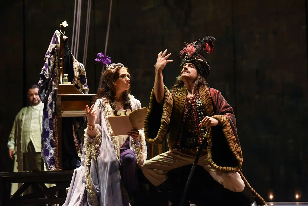 Escena de Adriana Lecouvreur con una mujer sentada leyendo un documento y un hombre con sombrero de plumas realizando un gesto teatral.