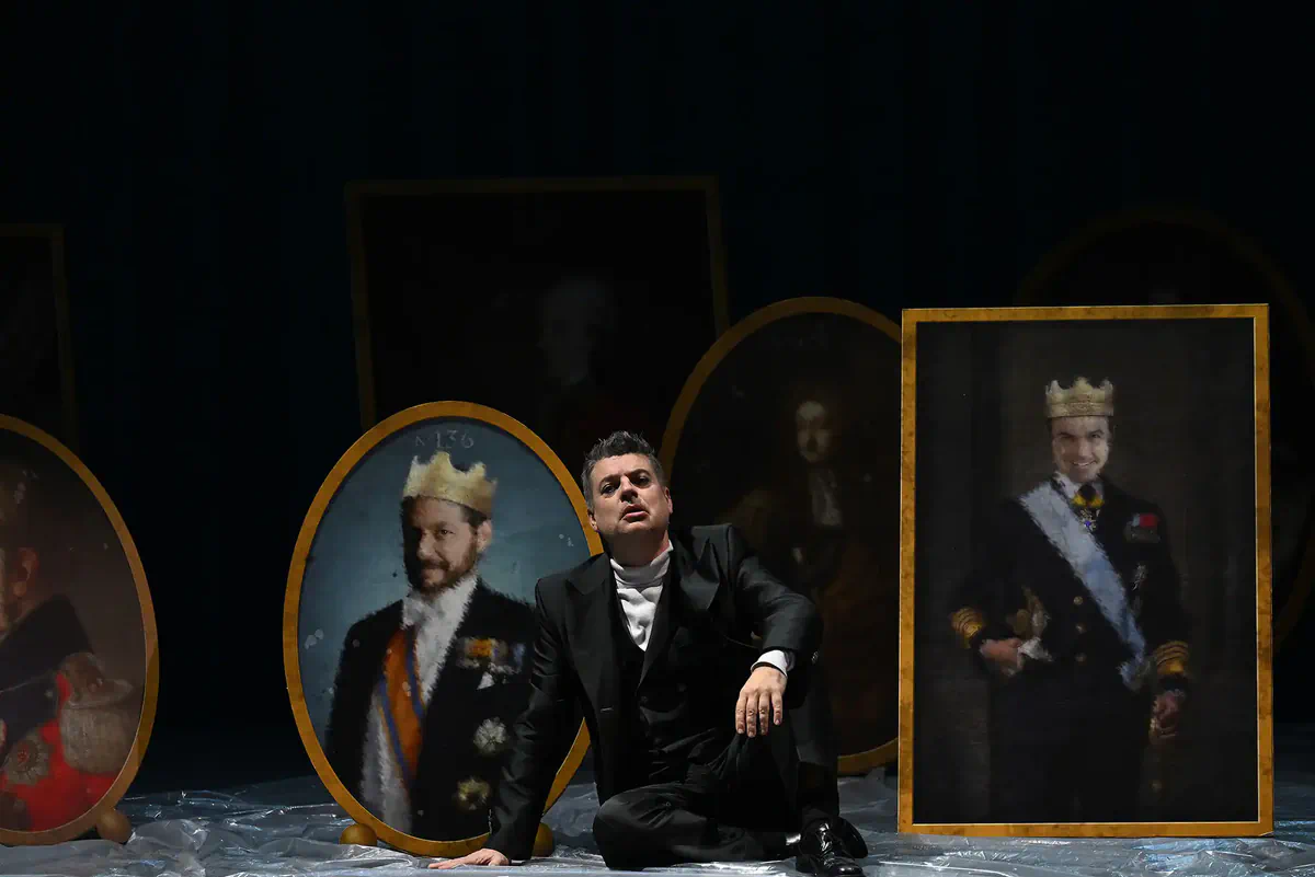 Escena de Hamlet con un intérprete sentado junto a varios retratos de personajes coronados.
