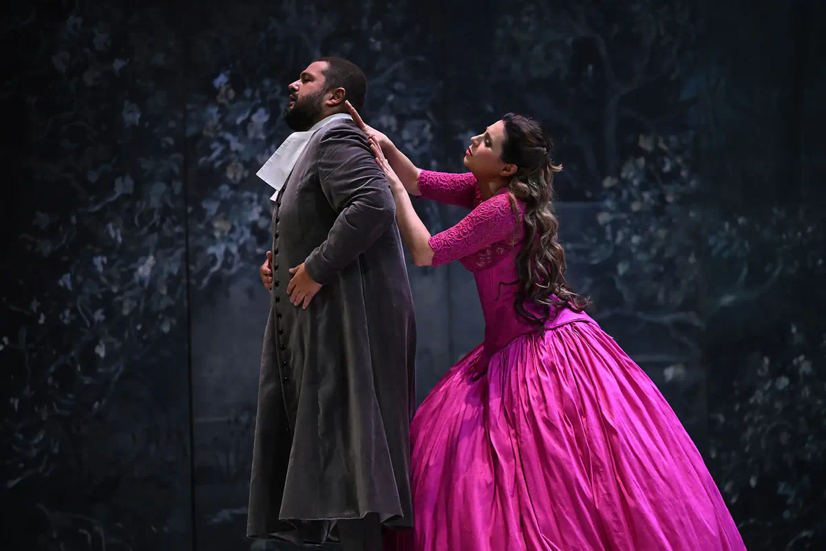 Escena de Manon con una mujer de vestido fucsia sujetando a un hombre por el cuello.